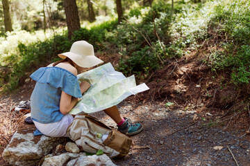 Woman hiker in wide‑brim hat studying a paper map while resting on a forest trail, outdoor navigation and route planning during a summer hike with backpack and travel gear