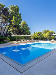 Luxurious hotel swimming pool area on a bright sunny day, featuring crystal-clear blue water, comfortable lounge chairs with umbrellas, and lush pine trees, perfect for showcasing summer vacations