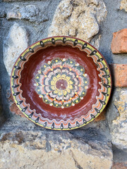 Vertical shot of a traditional hand-painted ceramic or clay plate with vibrant, colorful folk patterns, displayed on a rustic stone wall with visible brick accents