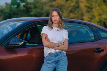 Young woman stands confidently next to her car in the evening light at a park