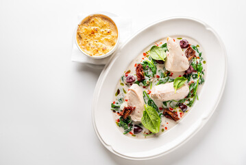 Creamy Tuscan chicken breast in a white sauce with spinach, sun-dried tomatoes, and olives on a white plate, with a side dish.
