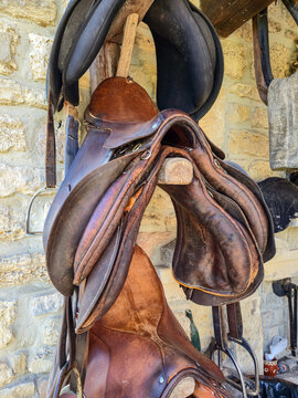 Rustic collection of worn, brown leather saddles and black tack hanging on a wooden rack against a rough, historic stone wall in a stable or rural tack room