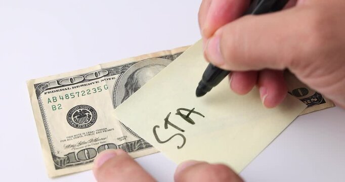 Hand writing the word STAKER on yellow post it beside 100 dollar bill on white background, concept of investment, finance and money management