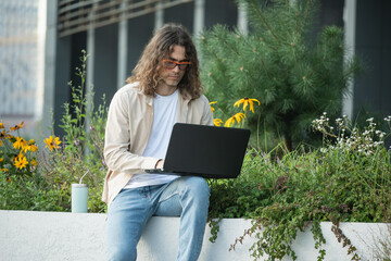 Handsome man, attractive freelancer, brunette sits on bench in urban park, using laptop for remote work. Young businessman or student enjoying work break Outdoors.