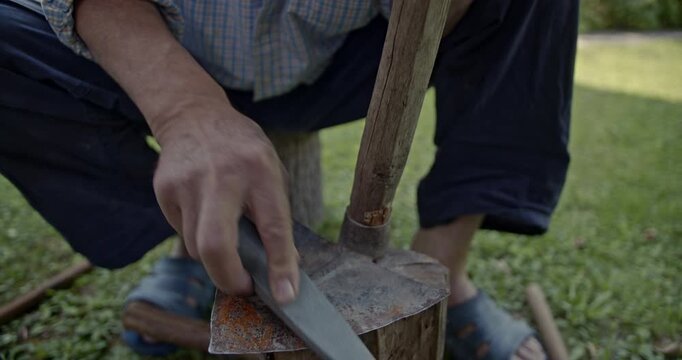 Watch a senior man diligently sharpen an aged gardening hoe or mattock using a traditional sharpening stone in the sunlight. The video includes authentic sounds of the process.
