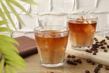 Tasty iced coffee with milk in glasses, beans and leaves on wooden table against light textured background, closeup
