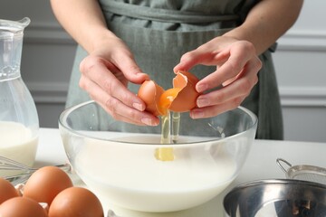 Making batter (liquid dough). Woman adding eggs into bowl at white table indoors, closeup