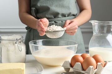 Making batter (liquid dough). Woman sieving flour into bowl at white table indoors, closeup