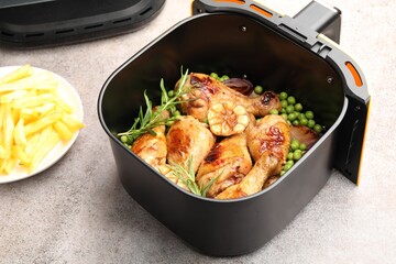 Air fryer basket with grilled chicken drumsticks, spices, green peas and french fries on grey table, closeup