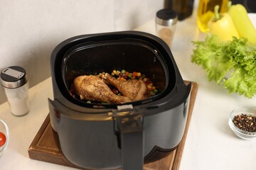 Air fryer basket with cooked chicken drumsticks, vegetables and spices on white marble countertop, closeup