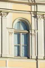 Close-up of an elegant arched window with decorative pilasters and ornate Corinthian capitals on a light yellow and white classical-style facade in sunlight.