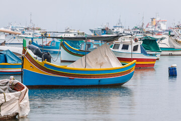 Fototapeta premium Colorful fishing boats are docked in a harbor on an overcast day, with many other vessels visible in the background.