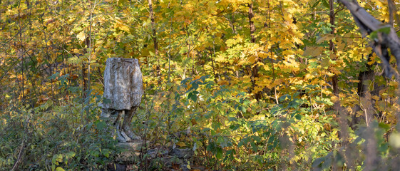 An old, weathered statue missing its head stands among dense foliage in a forest during autumn, surrounded by yellow and green leaves lit by soft sunlight.