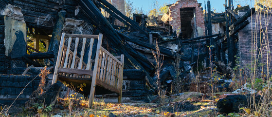 A wooden crib stands tilted amid the charred remains of a burned-down house, surrounded by overgrown vegetation and debris.