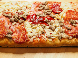 Close up meat feast pizza with different kind of salami and ham in rectangular shape on wooden background. Popular Italian dish with dough base, tomato sauce, cheese and other ingredients and spices.