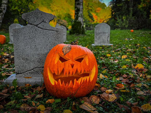 Scary Halloween pumpkin lantern with glowing face on autumn graveyard. Jack-o'-lantern in cemetery with fallen leaves and tombstones, spooky Halloween night atmosphere.