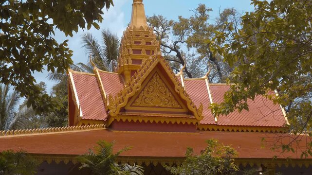Majestic tiled Buddhist temple roof with golden intricate gable and serpent-like chofas stand in the surroundings of trees crowns, Cambodia. Shot in motion