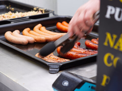 Hand using tongs to cook various sausages on black electric griddle pan in kitchen setting