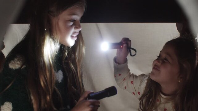 Girls having fun playing with flashlights inside a cozy fort at night