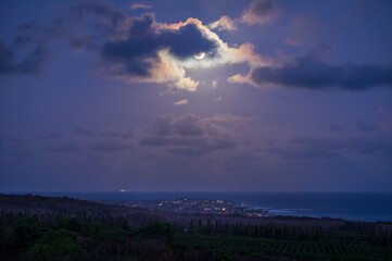 Dramatic moonrise over the Pacific Ocean and the resort town of Poipu, Kauai, Hawaii. The full moon peeks out from behind a cloud partially illuminating the coastline of this island paradise.
