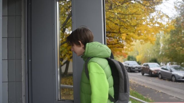 A 13 year old boy with a backpack opens the door of an apartment building in autumn.	