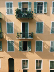 Fototapeta premium Warm yellow building with green shutters and balconies filled with plants in soft sunlight. Elegant Mediterranean architecture capturing charm, texture, and the relaxed lifestyle of southern Europe.