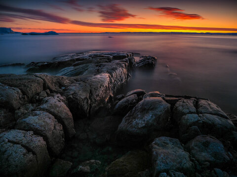 Moody Coastal Seascape at Sunrise or Sunset, Featuring Dark Foreground Rocks, Silky Water Effect (Long Exposure), and a Dramatic Orange and Red Horizon