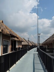 Scenic pathway connecting a series of charming bungalows with traditional thatched roofs under a bright, partly cloudy sky, evoking a serene tropical resort experience for a peaceful getaway