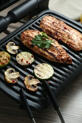 Electric grill with tasty chicken fillet, mushrooms, spices and zucchini on wooden table, closeup