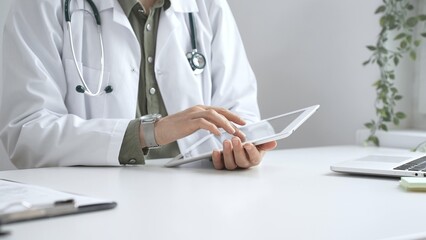Female doctor in lab coat and stethoscope using digital tablet at desk in medical office, accessing online patient information. Medicine and health care concept
