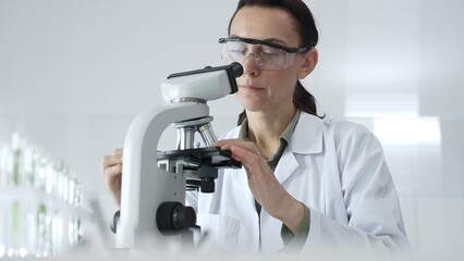 Female scientist in a lab coat and protective glasses examining samples through a microscope in a research laboratory. Medicine, healthcare and science concept