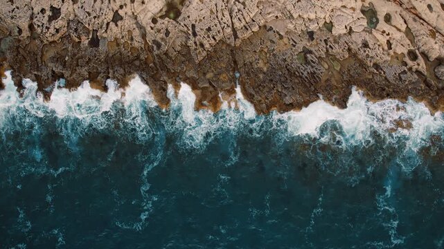 Aerial top down view of frothy ocean breaking over jagged brown stone shoreline