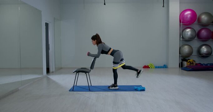 Athletic woman doing bent over glute kickback with chair in bright gym
