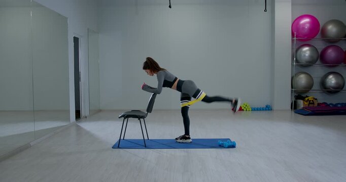 athletic woman is engaged in a bent over glute kickback while using a chair for support. She performs this exercise on a mat in a well lit gym focusing on strength training and fitness.
