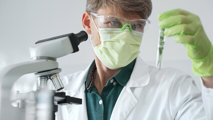 Adult male scientist wearing green mask, gloves and goggles using microscope and analyzing sample...