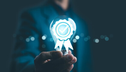 Businessman hand holding glowing light bulb with quality assurance certification badge and check...