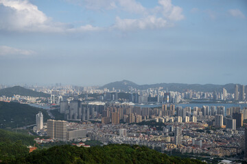 Obraz premium A Panoramic View of the Cityscape from a High Vantage Point Showing Dense Urban Buildings, Mountains, and a Coastal Water Body Under a Cloudy Sky