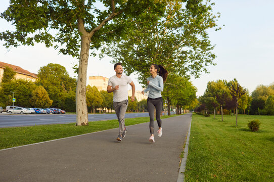 Sporty fit man and woman running together on paved street path, outdoor park trees and grass exercising and smiling, happiness and healthy lifestyle, connection and vitality enjoyed in active fitness - Powered by Adobe