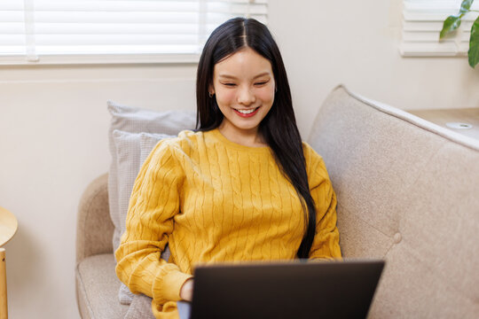 Photo of pretty young asian woman sit couch video call or work on laptop wear yellow pullover loft interior apartment indoors
