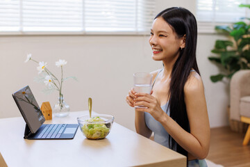 Happy young asian woman in Yoga outfit eating healthy salad while working with laptop at home office. Healthy lifestyle concept.
