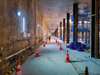 Underground construction hallway with steel support columns, safety cones, lighting, and scaffolding, showing active civil engineering and structural development work.