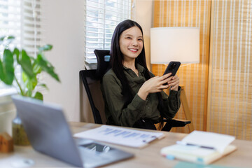 Serious charming asian woman using smartphone while working with laptop and financial documents laptop at home
