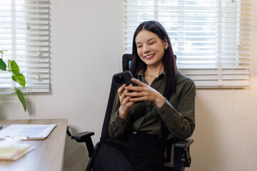 Serious charming asian woman using smartphone while working with laptop and financial documents laptop at home

