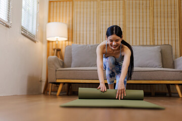 Female asian adult rolls yoga mat on wooden floor, suggesting preparation for exercise. Neutral...