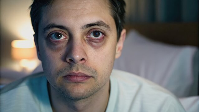 A tired man with dark circles around his eyes stares blankly, illuminated by soft light in the background, suggesting a mood of exhaustion or stress.