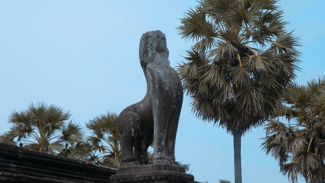 A ruined stone statue of a snow lion or singha is standing on a pedestal amongst palmyra palms in Angkor Wat, Cambodia. Shot in motion