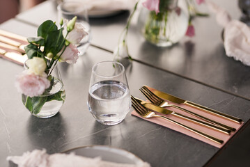 Elegant restaurant table close-up with gold cutlery, glass of water, and pastel flowers in small...