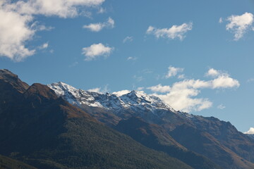Close-up of the rugged, snow-capped mountains of Glenorchy, New Zealand, on May 18, 2025. Lush green lower slopes meet dramatic peaks under a partly cloudy blue sky, showcasing the region's grandeur.