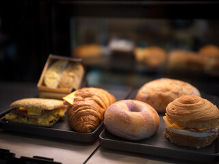 A variety of breakfast pastries displayed in a bakery, including croissants, bagels, sandwiches, and buns, arranged neatly on trays with a warm and inviting atmosphere