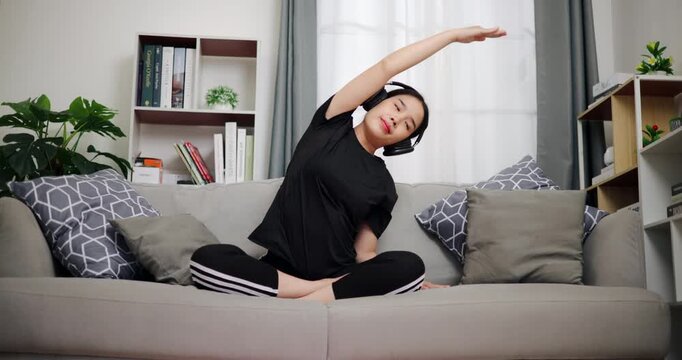 Handheld low angle view shot, Relaxed Asian woman is practicing yoga on the sofa, enjoying music through wireless headphones in a cozy room at home. Women Lifestyle